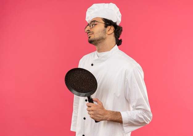 With closed eyes young male cook wearing chef uniform and glasses holding frying pan on pink