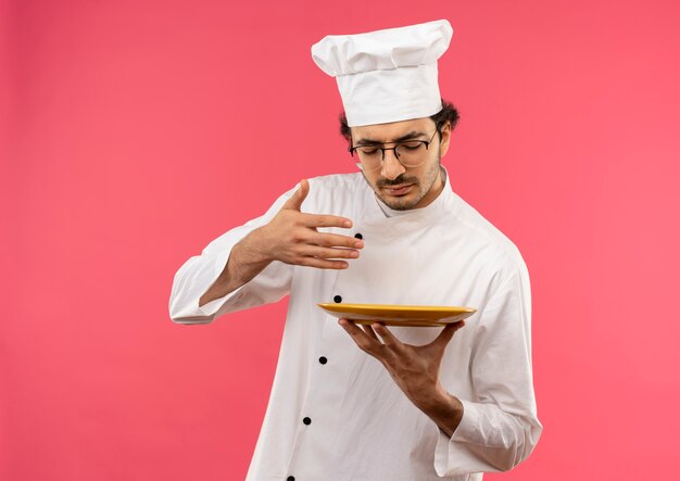 Con gli occhi chiusi giovane cuoco maschio che indossa l'uniforme dello chef e occhiali fingendo pasto sniffing sul cibo isolato sulla parete rosa