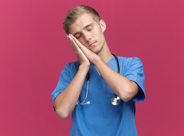 With closed eyes young male doctor wearing doctor uniform with stethoscope showing sleep gesture isolated on pink wall with copy space