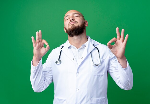 С закрытыми глазами молодой врач-мужчина в медицинском халате и стетоскопе показывает жест, изолированный на зеленом