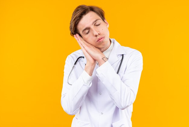 With closed eyes young male doctor wearing medical robe with stethoscope showing sleep gesture isolated on orange wall