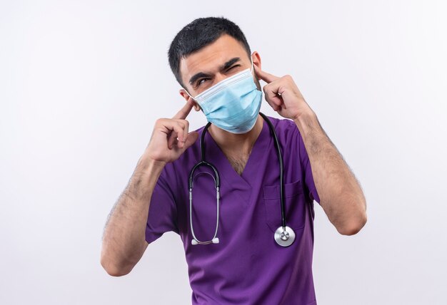 С закрытыми глазами молодой врач-мужчина в фиолетовой одежде хирурга и медицинской маске со стетоскопом закрыл уши на изолированном белом