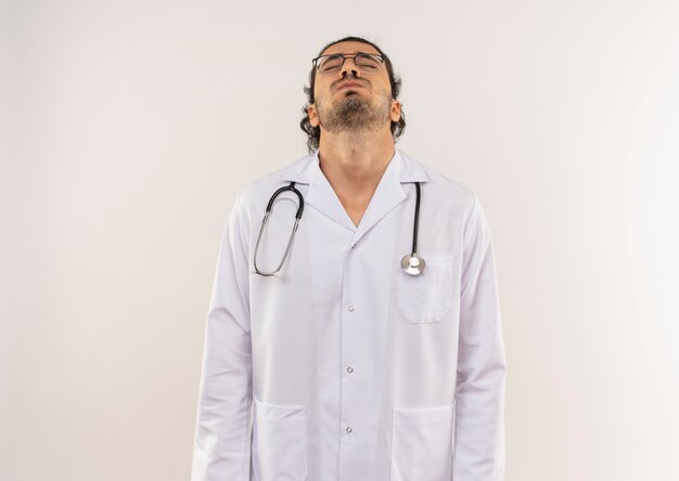 With closed eyes young male doctor with optical glasses wearing white robe with stethoscope raising head on isolated white wall with copy space