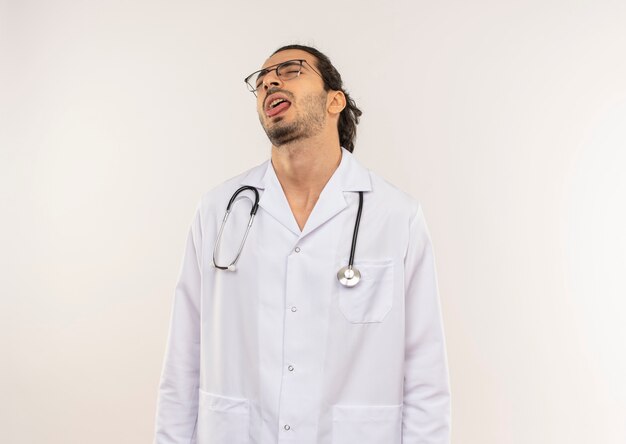 With closed eyes young male doctor with optical glasses wearing white robe with stethoscope showing tongue on isolated white wall with copy space