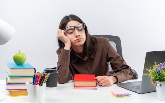 With closed eyes young school woman wearing glasses sits at table with school tools 