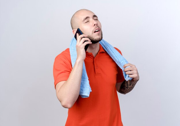 With closed eyes young sporty man speaks on phone with towel on shoulder isolated on white