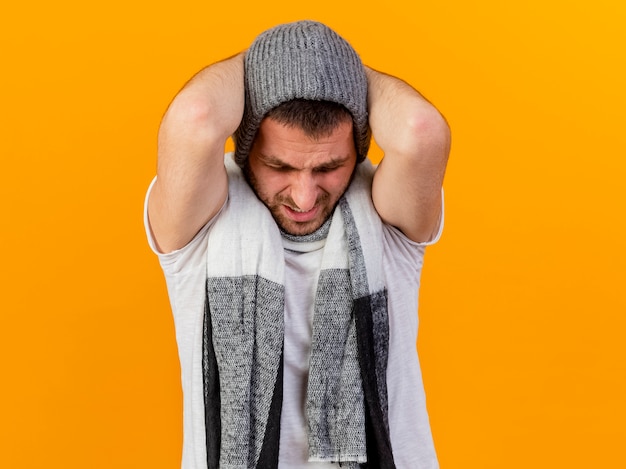 With lowered head regretted young ill man wearing winter hat and scarf grabbed head isolated on yellow background