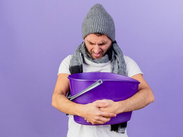 With lowered head unpleased young ill man wearing winter hat with scarf holding plastic bucket and vomiting into it isolated on purple background