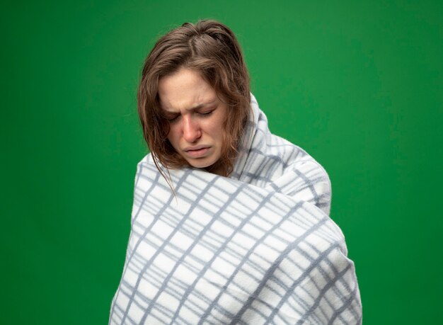 With lowered head weak young ill girl wearing white robe wrapped in plaid isolated on green