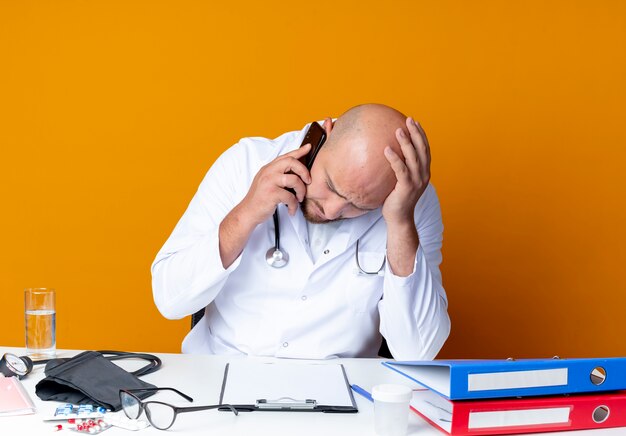 With lowered head young bald male doctor wearing medical robe and stethoscope sitting at work desk with medical tools speaks on phone on orange