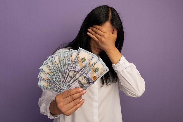 With lowered head young beautiful girl wearing white t-shirt holding cash isolated on purple