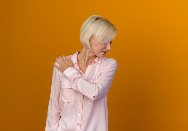 Foto gratuita con la testa abbassata giovane donna bionda slava che mette la mano sulla spalla isolata sull'arancio con lo spazio della copia