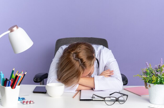 Con la testa abbassata, una giovane dottoressa che indossa una tunica medica con uno stetoscopio si siede al tavolo con strumenti medici isolati su sfondo blu