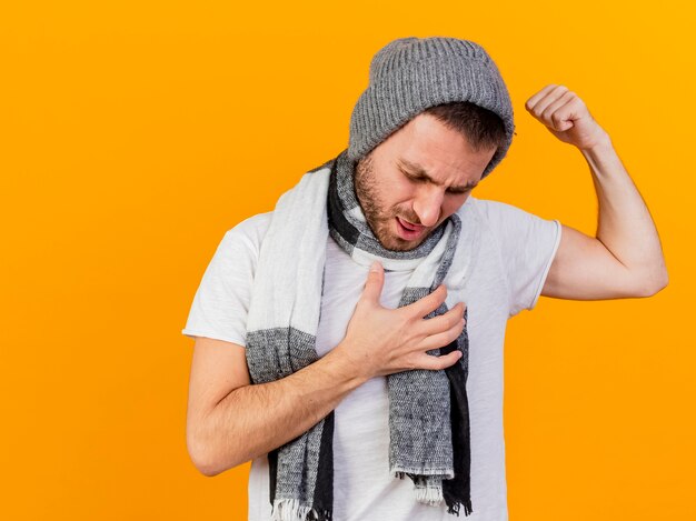 Con la testa abbassata giovane uomo malato che indossa cappello invernale e sciarpa che mostra un forte gesto mettendo la mano sul cuore isolato su sfondo giallo