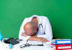 Free photo with lowered head young male doctor wearing medical robe and stethoscope sitting at desk with medical tools isolated on green background