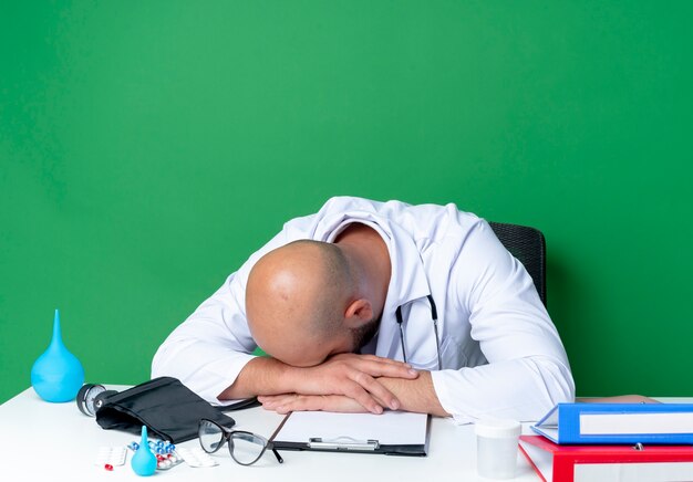 With lowered head young male doctor wearing medical robe and stethoscope sitting at desk with medical tools isolated on green wall