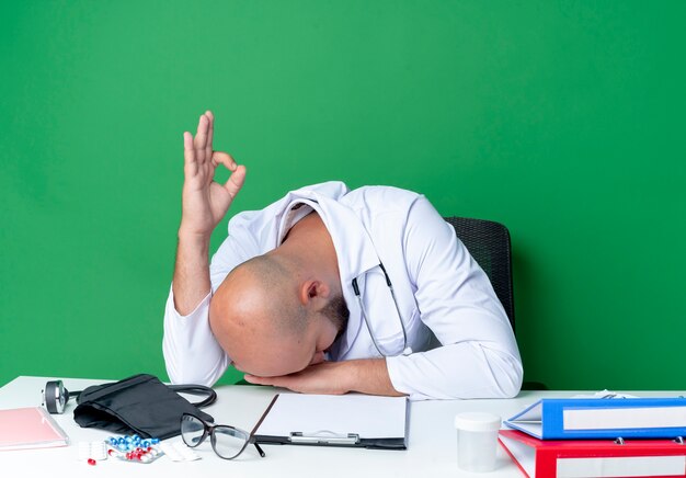 With lowered head young male doctor wearing medical robe and stethoscope sitting at desk