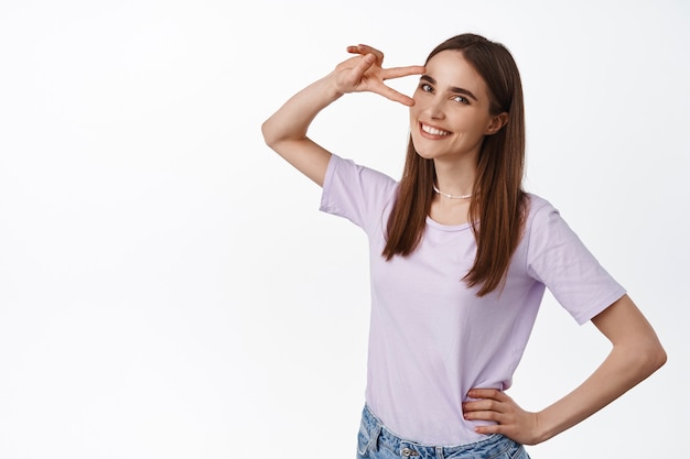 woman 25s years, showing peace disco v-sign near eye, smiling and looking lovely , standing on white