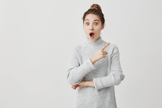 Woman 30s standing with open mouth and showing index finger at something exciting. Funny emotions of female student being shocked and excited. Amusement concept