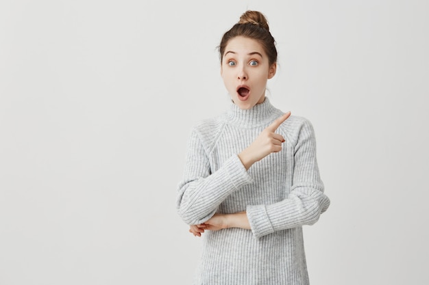 Woman 30s standing with open mouth and showing index finger at something exciting. Funny emotions of female student being shocked and excited. Amusement concept