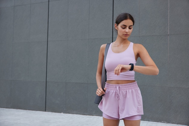 woman in activewear checks results of fitness training on wearable wristwatch carries mat poses against grey wall with copy space