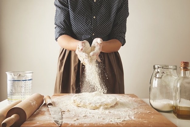 Женщина добавляет немного муки в тесто на деревянном столе возле ножа, две скалки, мерный стаканчик, прозрачную бутылку с мукой и оливковым маслом. Пошаговое руководство по приготовлению макаронных пельменей