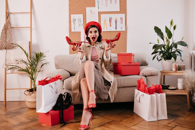 Woman after shopping sitting on sofa with new apparel. Beautiful fashionable girl holds red modern shoes and sits on sofa.