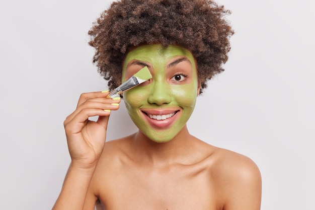 woman applies green homemade natural mask with cosmetic brush smiles toothily stands topless has healthy skin isolated on white 