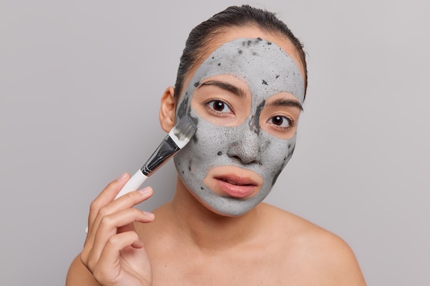 woman applies purifying clay mask on face with cosmetic brush enjoys skin care treatment looks directly at camera models naked on grey 