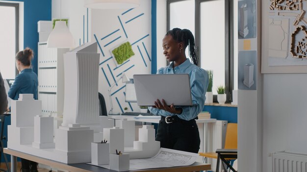 건축 사무실에서 건물 모델을 분석하기 위해 노트북을 들고 있는 여성 건축가. 도시 프로젝트를 위한 건설 구조를 설계하기 위해 컴퓨터와 마케트로 작업하는 엔지니어.