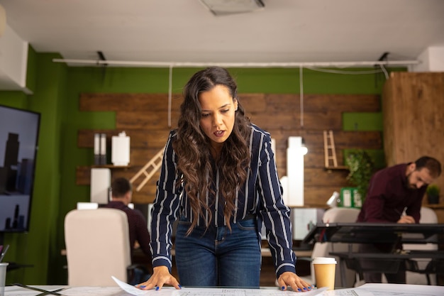 Woman architect looking in blue print on table. Colleagues working in the back