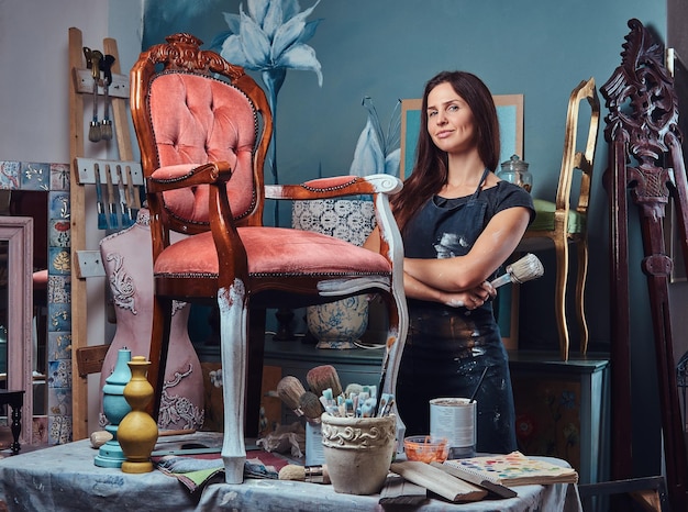 Woman artist in apron holds paintbrush while standing with crossed arms in workshop.