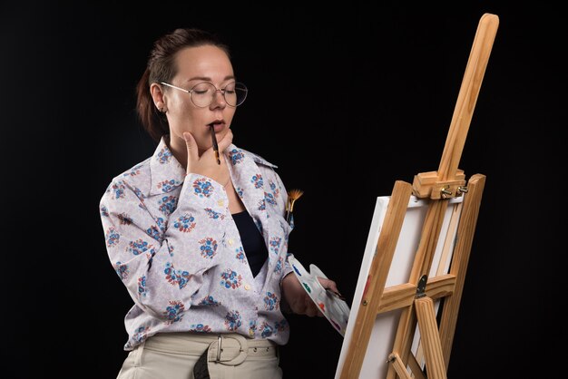 Woman artist holding brush and looking at canvas on black background