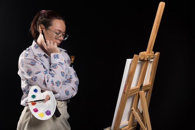 Woman artist holding brush and looking at canvas on black background