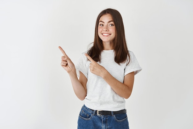 Woman asking to check this out, pointing fingers at upper left corner promo offer and smiling, showing advertisement on white wall.