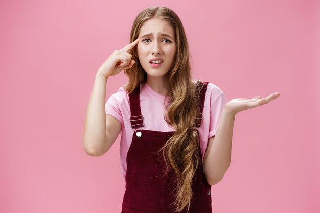 Woman asks if friend stupid or weird. Questioned displeased and bothered girl with long wavy natural hairstyle and pure clean skin shrugging with raised palm rolling finger over temple with contempt.