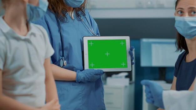 オフィスでタブレットに水平方向の緑色の画面を保持している女性アシスタント。検診時にガジェットディスプレイにクロマキーモックアップと空白の背景を持つ分離コピースペーステンプレートを使用している医療看護師。