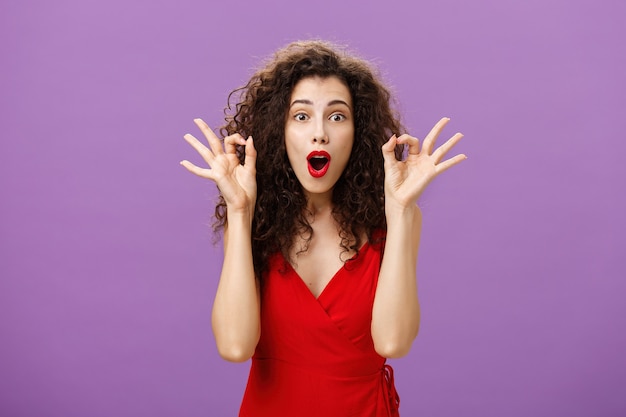 Woman assring husband he doing great supporting him during poket match standing amazed and excited over purple wall in attractive red dress showing okay or excellent sign folding lips from excitement.