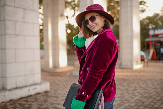 Woman in autumn style trendy outfit walking in street wearing purple velvet jacket, sunglasses and hat
