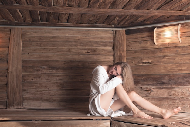 Woman in bathrobe sitting on wooden bench relaxing at sauna