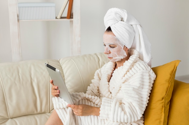 Женщина в халате с маской для лица, используя свой планшет