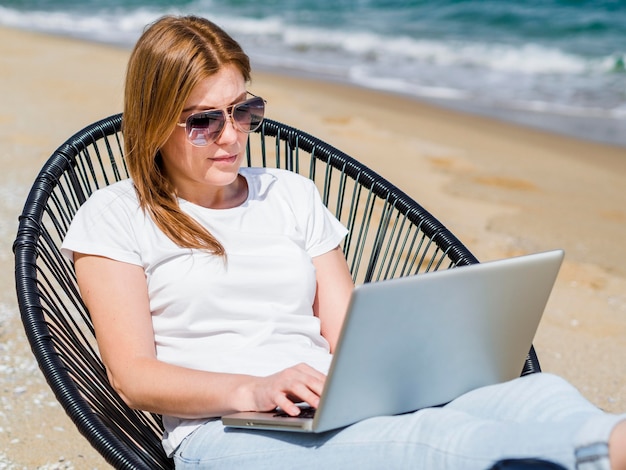 Donna nella sedia di spiaggia che lavora al computer portatile mentre indossando gli occhiali da sole