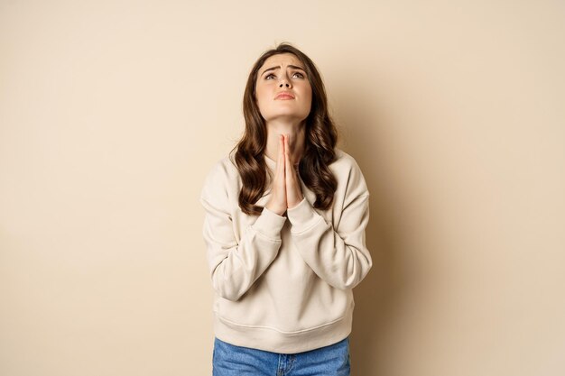 Woman begging and pleading god, supplicating, say please, standing desperate over beige background