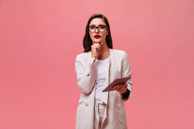 Woman in beige suit poses thoughtfully and holds tablet. Serious girl in light fashionable outfit and glasses poses for camera.