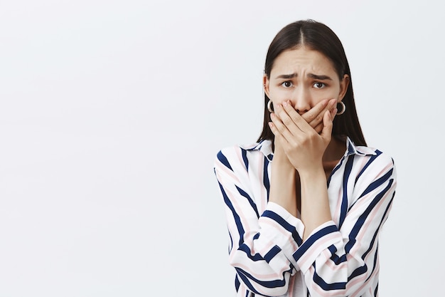 Woman being harassed afraid to tell anyone. Scared anxious woman in striped blouse and trendy earrings, covering mouth with palms not to scream, frowning, being afraid over gray wall