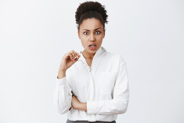 Woman being shocked listening to terrible news on tv. Portrait of shook displeased and questioned woman in shirt, biting rim of glasses staring with popped eyes and frowning from stupor