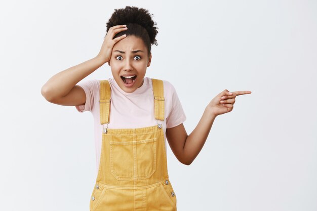 Woman being stunned with ridiculous prices on food in cafe. Shocked and displeased dark-skinned female student in trendy yellow dungarees, holding palm on head, talking with shock and pointing right