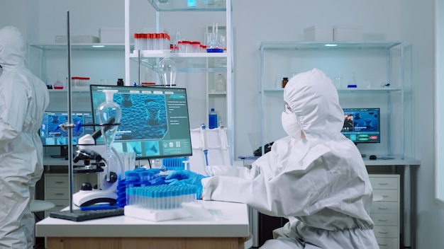 Woman biochemist in coverall checking manifestations of virus working on comuper in equipped lab. Team of doctors examining vaccine evolution using high tech researching diagnosis against covid19