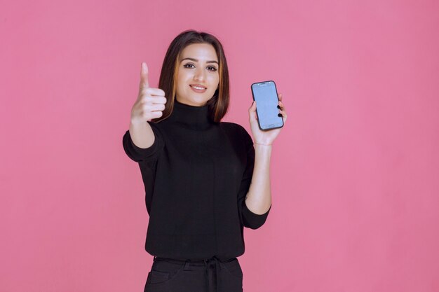 Женщина в черном свитере держит смартфон и делает большой палец вверх.