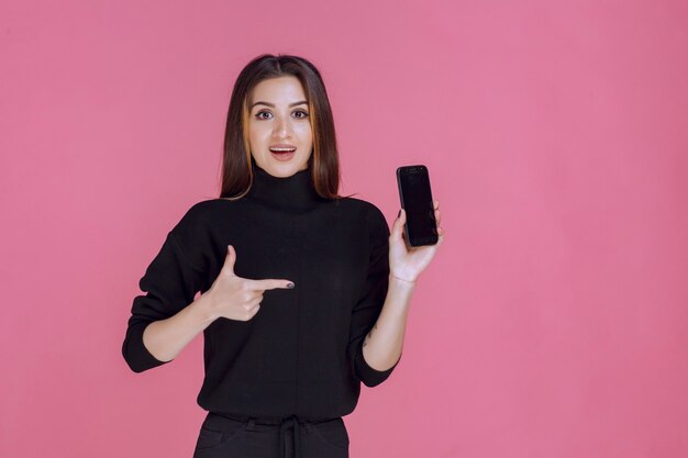 Woman in black sweater holding a smartphone and pointing at it. 